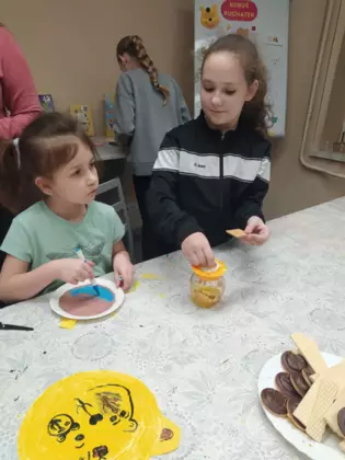 Dziewczynka maluje mazakami obrazek a druga smaruje miodem ciasteczko.
