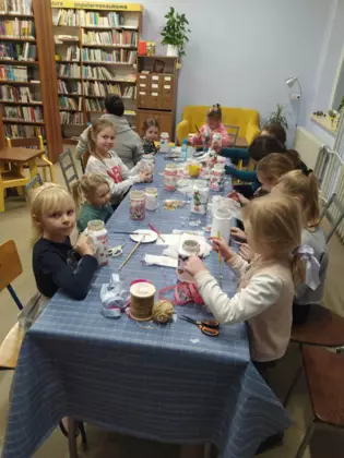 Uczestnicy siedzą przy stole w bibliotece i wykonują prezenty na Dzień Babci i Dziadka