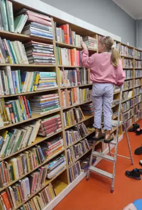 Dziewczynka stoi na drabinie poszukując odpowiedzi na zagadkę w zbiorach bibliotecznych.