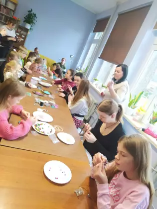 Uczestnicy zajęć siedzą przy stole i wykonują zestawy biżuterii.