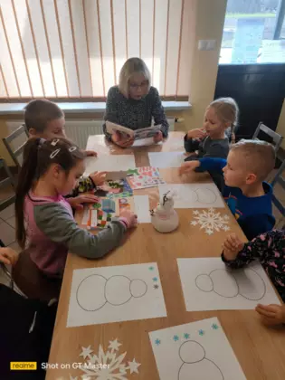 Przedszkolaki siedzą przy stolikach, słuchają czytanej bajeczki i kolorują bałwanki.