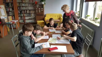 Dzieci siedzą w bibliotece przy stoliku i z pmocą pani bibliotekarki rozwiązują krzyżówki ortograficzne.