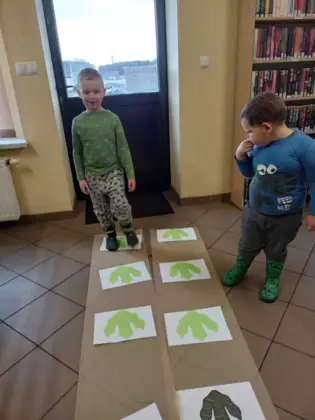 Dwóch chłopców chodzi po śladach dinozaurów rozłożonych na podłodze  w bibliotece.