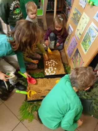 Przedszkolaki wykopują z piasku gipsowe figurki dinozaurów.