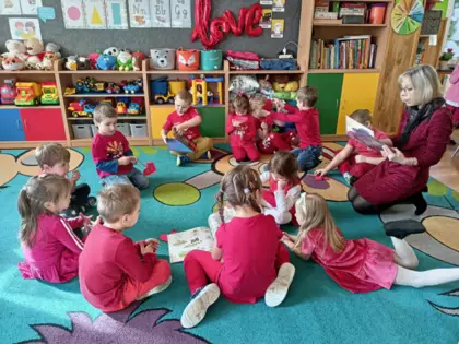 Przedszkolaki siedzą na dywanie i słuchają bajki czytanej przez bibliotekarkę.