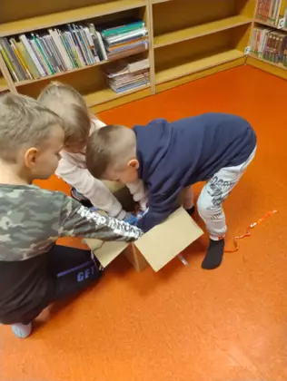 Dwóch chłopców i dziewczynka przeszukują karton w poszukiwaniu rozwiązania postawionego przed nimi zadania.