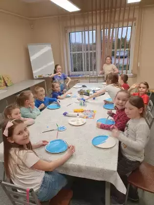 Dzieci siedzą przy stole i malują talerzyki papierowe niebieską farbą.
