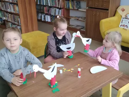 Dzieci siedzą przy stoliku trzymając bociany z papieru w ręce.