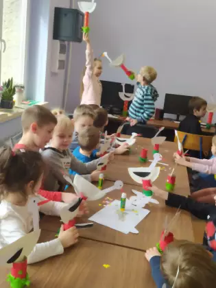 Dzieci siedzą przy stoliku i wykonują bociany z papieru.