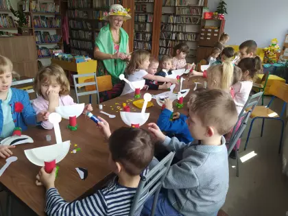 Dzieci siedzą w bibliotece przy stoliku i kleją bociany z papieru.