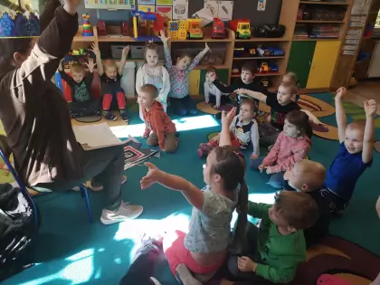 Dzieci w przedszkolu siedzą na dywanie i podnoszą radośnie ręce do góry