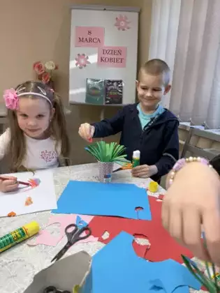 Dziewczynka koloruje kredkami a chłopczyk przykleja kwiatki do trawy z papieru.