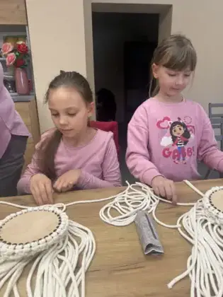 Dwie dziewczynki splatają sznurki makramy tworząc koszyczek.