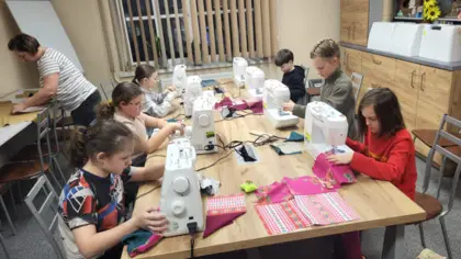 Szóstka dzieci siedzi przy stole i szyją na maszynach. Z lewej strony pani wycina materiał.