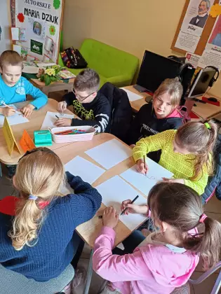 Uczniowie siedzą przy stole i kolorują obrazki.