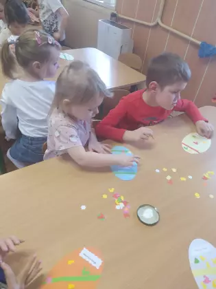 Przedszkolaki siedzą przy stoliku i kleją ozdoby wielkanocne.
