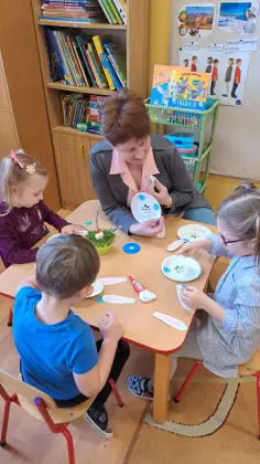 Dzieci siedzą przy stoliku z Panią bibliotekarką i kolorują pisanki wielkanocne.