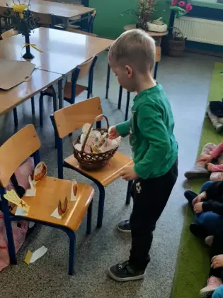 Chłopczyk wkłada do koszyka wielkanocnego pokarm wycięty z papieru.