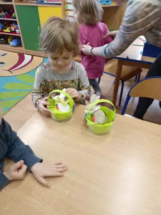 Dziewczynka siedzi przy stoliku i wkłada do koszyczka gipsowego zajączka.