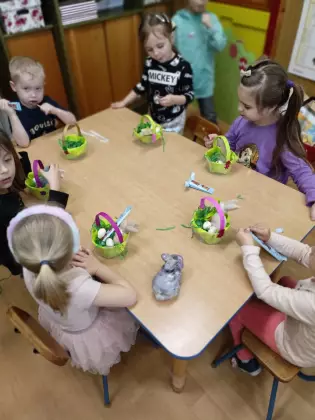 Dzieci siedzą przy stoliku w przedszkolu. Na stole stoją koszyczki wielkanocne.