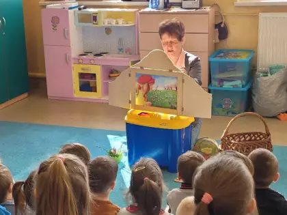 Dzieci siedzą na dywanie i słuchają opowiadania przedstawianego w formie teatrzyku kamishibai.