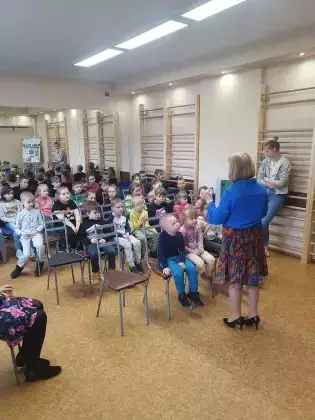 Dzieci siedzą na krzesełkach w sali i słuchają czytanego opowiadania.