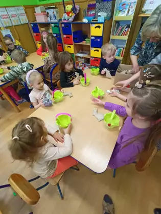 Dzieci siedzą przy stole i wyklejają koszyczki wielkanocne.