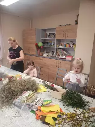 Uczestnicy warsztatów siedzą przy stole i owijają drewniane kółko sianem.