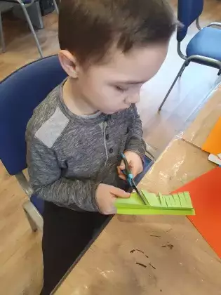 Chłopczyk siedzi przy stoliku i wycina z papieru zwierzątko.