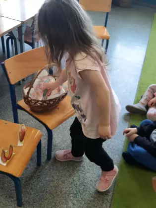 Dziewczynka wkłada do koszyka wielkanocnego pokarm wycięty z papieru.