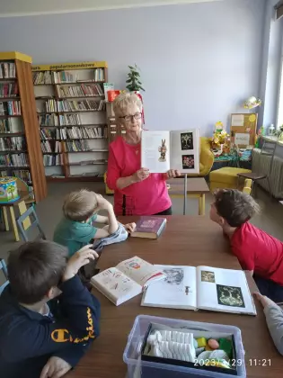 dzieci siedzą przy stole i słuchają opowiadania czytanego przez bibliotekarkę.