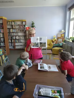 Dzieci siedzą przy stole w bibliotece i słuchają opowiadania.