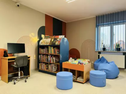 Strefa Młodego Czytelnika w bibliotece na środku stoi regał z książkami obok znajduje się biurko z komputerem oraz pufki do siedzenia.