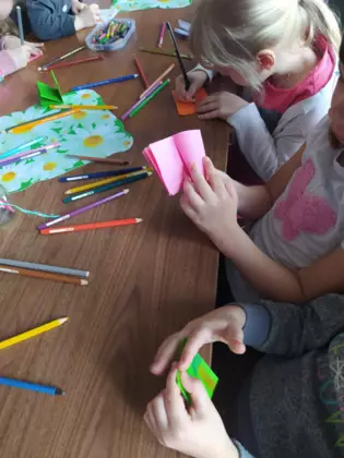 Przedszkolaki siedzą przy stole w bibliotece i malują obrazki w mini książeczkach.