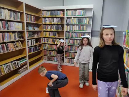 Dzieci chodzą w bibliotece z książką na głowie.