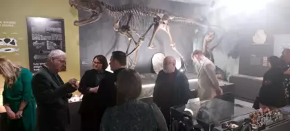 Uczestnicy otwarcia muzeum oglądają eksponaty. Za nimi stoi rekonstrukcja szkieletu dinozaura.