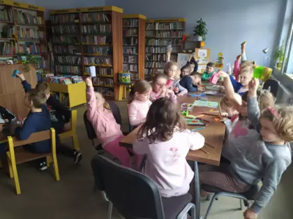 Przedszkolaki siedzą przy stole w bibliotece i malują obrazki w mini książeczkach.