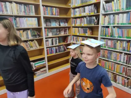 Dzieci chodzą w bibliotece z książką na głowie.