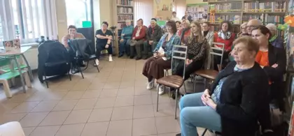 Uczestnicy spotkania autorskiego siedzą w bibliotece i słuchają prelekcji.