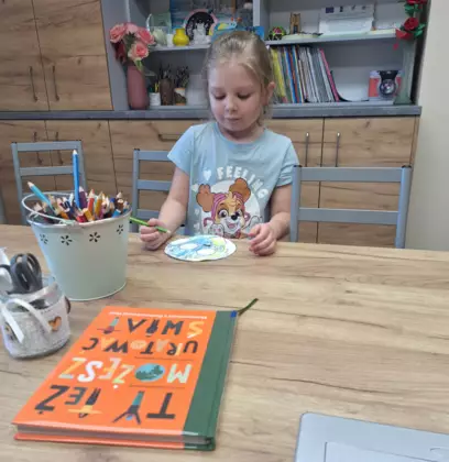 Uczestniczka zajęć koloruje kredkami Ziemię. Przed nią na stole leży książka.