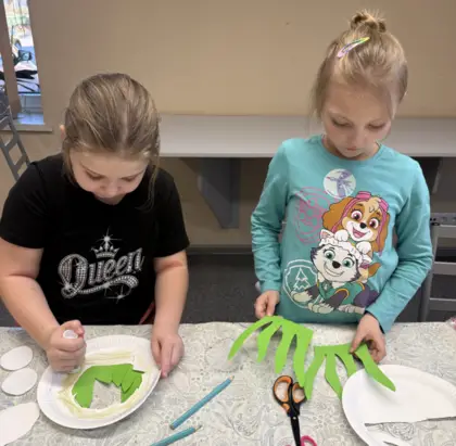 Dwie dziewczyny wycinają trawę z papieru kolorowego i przyklejają do talerzyka papierowego.