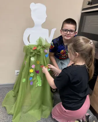 Dziewczynka przykleja ozdoby do sukni pani wiosny a chłopczyk w okularach uśmiecha się do  niej.