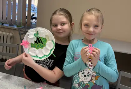 Dwie uśmiechnięte dziewczyny. Jedna trzyma w rękach przebiśniegi przyklejone na talerzyku papierowym.