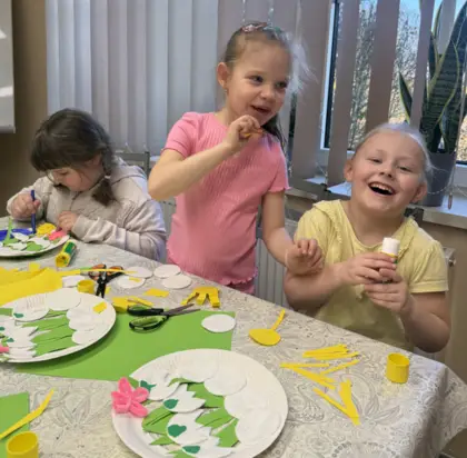 Dwie uśmiechnięte dziewczyny przyklejają kolorowy papier na talerzyk papierowy. Trzecia dziewczyna w skupieniu wykonuje swoją prace.