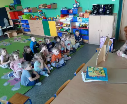 Przedszkolaki siedzą na dywanie i słuchają opowiadania czytanego w formie teatrzyku.