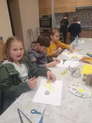 Dzieci siedzą przy stole w bibliotece i malują farbami pszczoły z tektury.