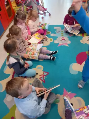 Przedszkolaki siedzą na dywanie i oglądają książki przyniesione z biblioteki.