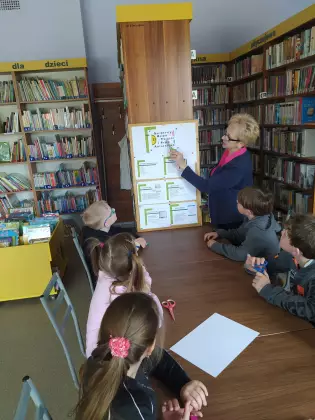 Uczniowie w bibliotece słuchają ciekawostek na temat książki opowiadanej przez Panią bibliotekarkę.