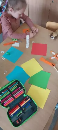 Dziewczynka siedzi przy stoliku i klei z papieru zakładkę do książki.