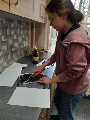 Uczestniczka zajęć maluje wałkiem z czarną farbą kartkę papieru.